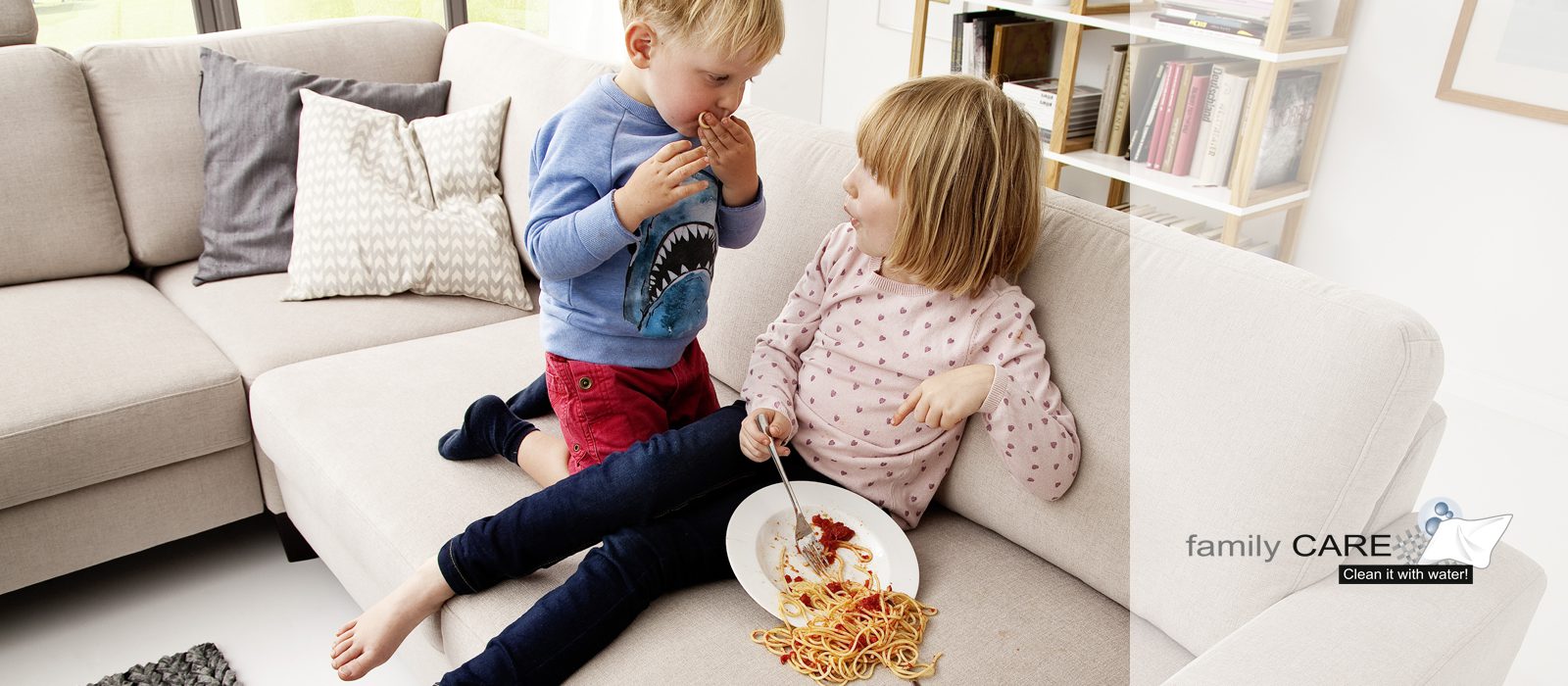 Family Care - Zwei Kinder auf der Couch mit einem Teller Nudeln und Tomatensoße, der auf die Couch geleert ist.
